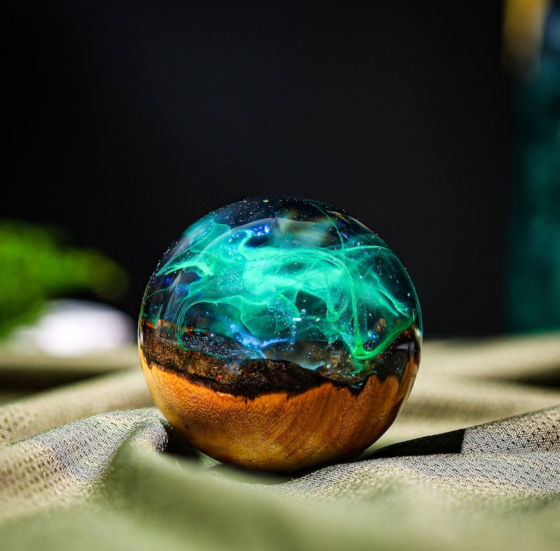 May include: A round, wooden orb with a clear resin top filled with swirling green and blue resin. The resin looks like a galaxy or nebula.