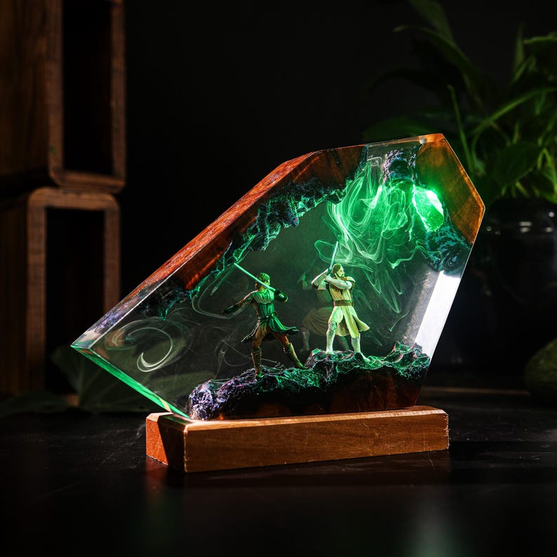 May include: A unique resin sculpture featuring two figures with glowing green lightsabers, set within a clear, angled block. The sculpture has a wooden base and a dark background, with green and white swirling patterns inside the resin.