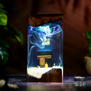 May include: A rectangular resin and wood block lamp featuring a miniature black Kaaba structure at the base. The clear resin section contains swirling blue and purple smoke-like patterns with the golden Arabic word Allah floating above the Kaaba.