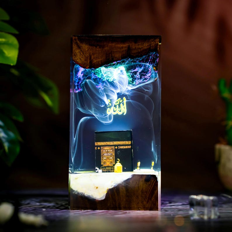 May include: A rectangular resin and wood block lamp featuring a miniature black Kaaba structure at the base. The clear resin section contains swirling blue and purple smoke-like patterns with the golden Arabic word Allah floating above the Kaaba.