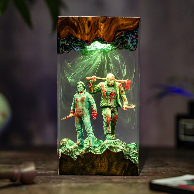 May include: A rectangular resin art piece featuring two horror movie figures, one holding an axe and the other a knife. The figures are green with red accents, set against a smoky green background. The top is wood-grained, and the base is textured green.