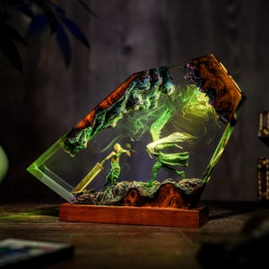 May include: A unique resin art piece featuring a battle scene with two figures, one with a sword. The artwork is encased in a clear, angled block with a wooden base. The scene is illuminated with green and yellow light.