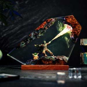 May include: A decorative resin and wood lamp featuring a miniature armored figure in a cape extending an arm to emit a glowing yellow light beam toward a rocky formation, with a small green creature standing nearby on a dark wooden base.