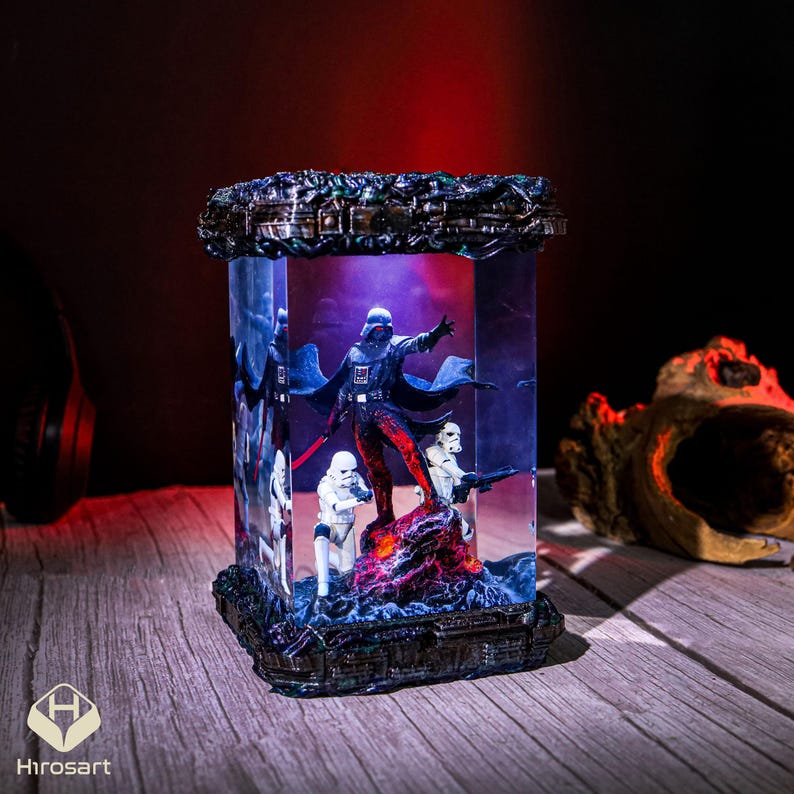 K&ouml;nnte beinhalten: Ein klarer Glasw&uuml;rfel mit einem schwarzen Metallrahmen umschlie&szlig;t eine Miniatur-Szene mit Darth Vader und Stormtroopers. Die Szene wird von unten mit blauem Licht beleuchtet, was einen dramatischen Effekt erzeugt.