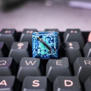 May include: A blue and green keyboard keycap with a stylized image of a sword inside. The keycap is made of resin and has a textured surface.