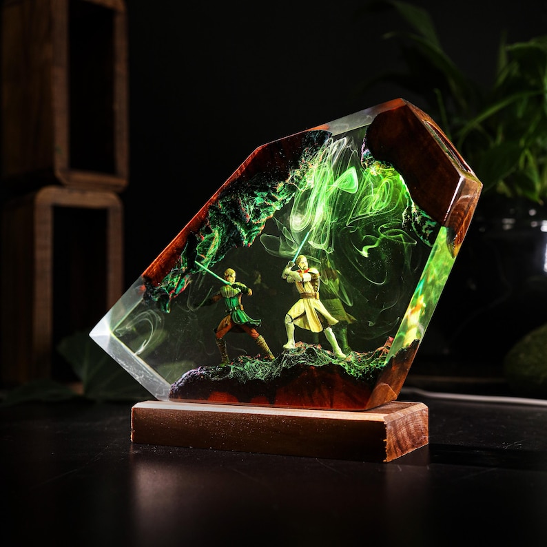 May include: A decorative resin sculpture featuring two figures with glowing green lightsabers in a battle scene. The sculpture is encased in clear resin with a wooden base and accents, creating a 3D effect.