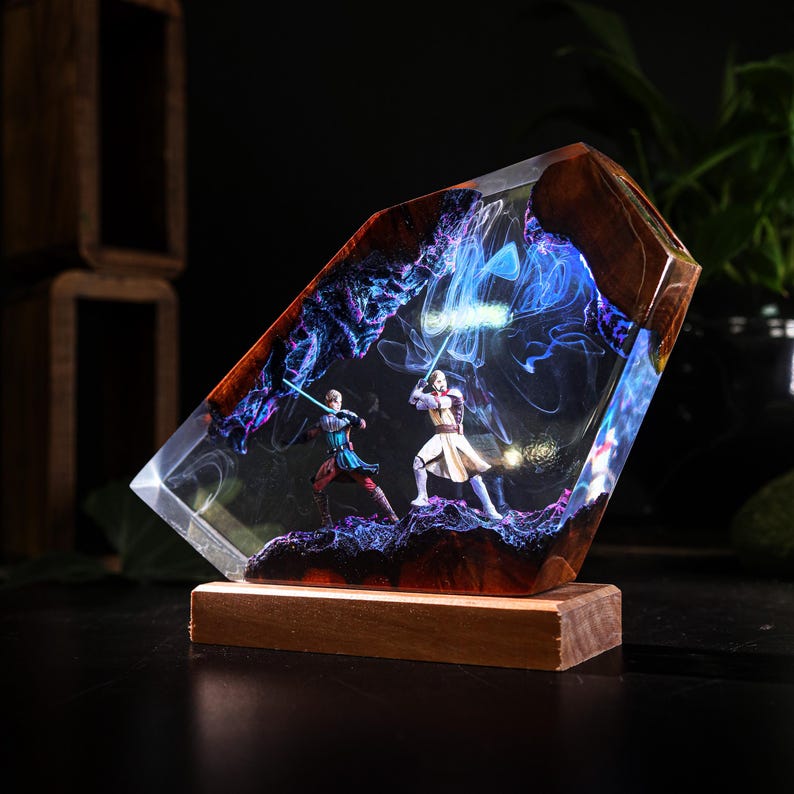 May include: A resin sculpture featuring two figures with lightsabers in a battle scene. The figures are set within a clear resin block with a wooden base. The scene is illuminated with blue and purple light effects.