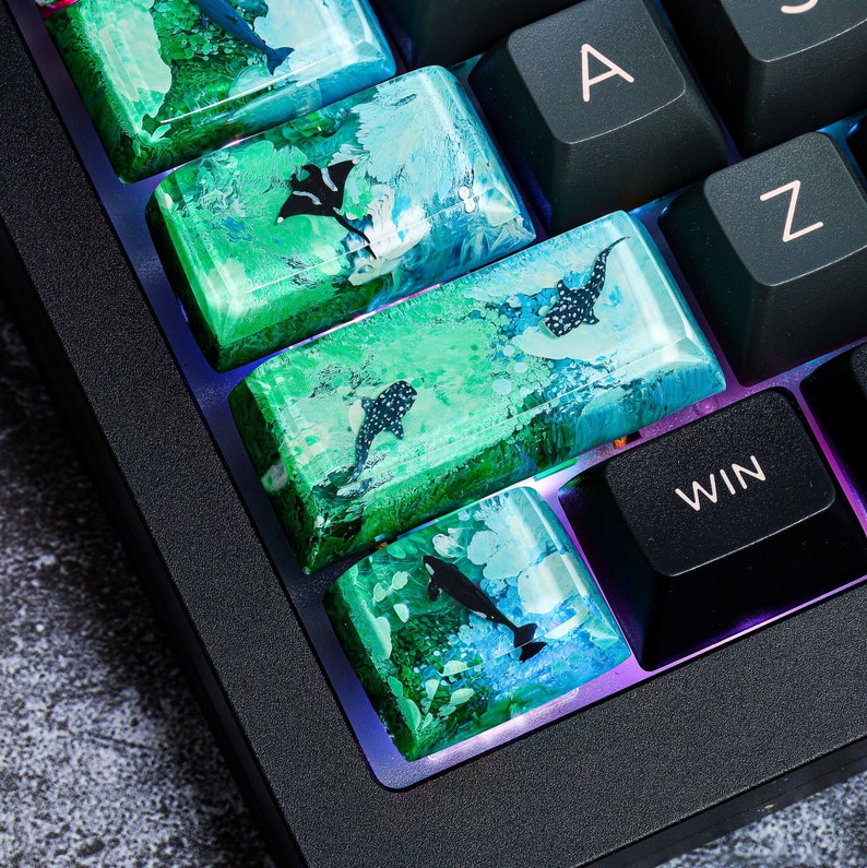May include: A close-up of a keyboard with custom keycaps featuring a green and blue ocean-themed design with black silhouettes of sharks and rays. The keycaps are made of resin and have a glossy finish. The keyboard has a black frame and black keys with white lettering. The keys labeled "A", "Z", and "WIN" are visible.