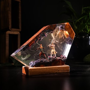 May include: A decorative resin sculpture featuring two figures with lightsabers in a combat pose. The figures are set within a clear resin block with a wooden base. The resin has a swirling, smoky effect with purple and orange accents.