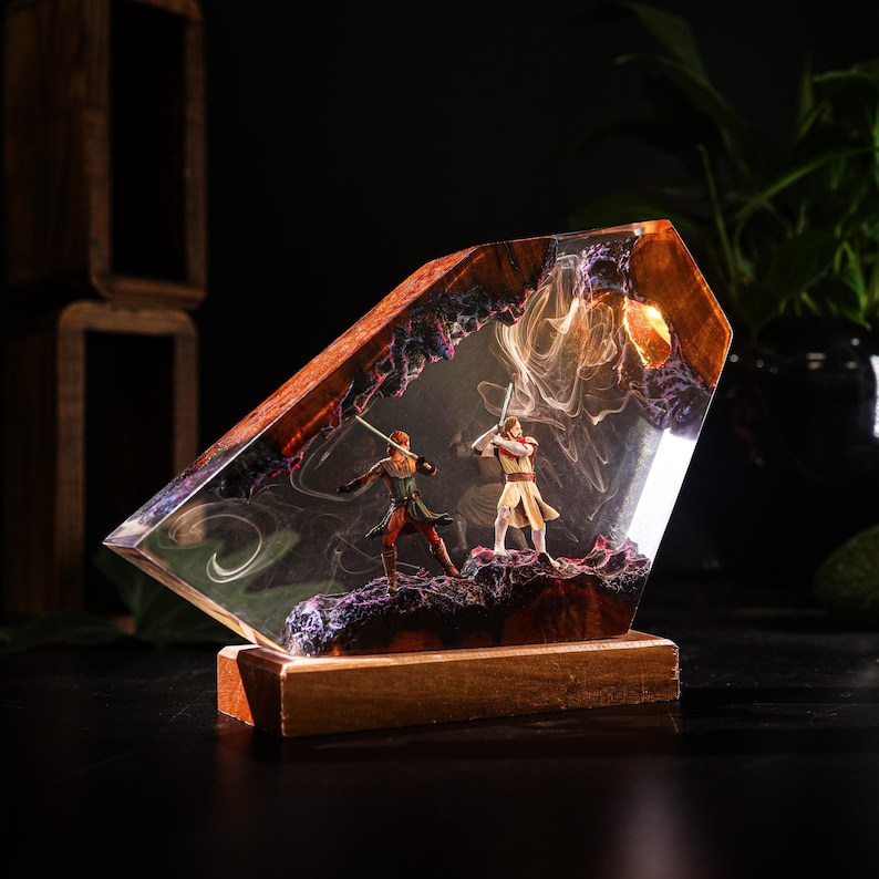 May include: A decorative resin sculpture featuring two figures with lightsabers in a combat pose. The figures are set within a clear resin block with a wooden base. The resin has a swirling, smoky effect with purple and orange accents.