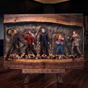 May include: A rectangular live-edge wood board featuring a central resin inlay containing five horror movie character action figures standing on a bed of miniature skulls. The figures include a masked slasher, a striped sweater character, a man in a jumpsuit, a doll with a bubblegum, and a clown.