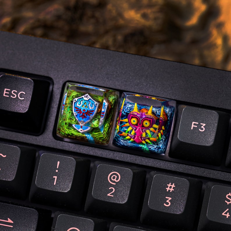 May include: Two custom keyboard keycaps featuring characters from the Legend of Zelda video game series. The left keycap is green and blue with the Triforce symbol and the right keycap is blue and pink with the Majora's Mask.