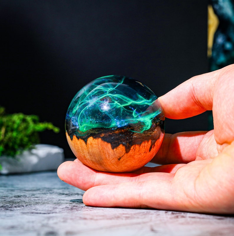 May include: A round, wooden orb with a clear resin top filled with swirling green and blue glitter. The orb is about the size of a baseball and is being held in a hand.