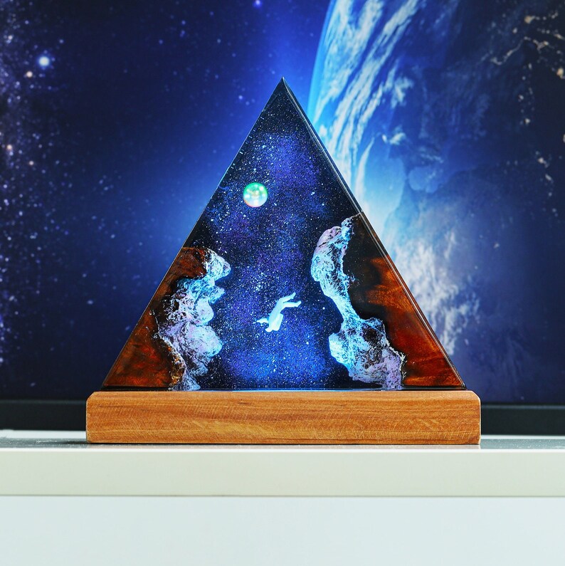 May include: A triangular resin pyramid with a blue, black, and white galaxy design. The pyramid is lit from within, creating a glowing effect. The base of the pyramid is made of wood.