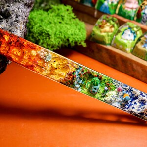 May include: A long, clear, rectangular resin bar with a rainbow of colors and embedded flowers and other botanical elements. The bar is on a brown surface.