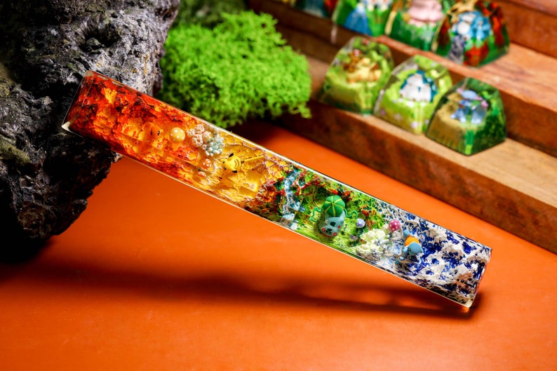 May include: A long, clear, rectangular resin bar with a rainbow of colors and embedded flowers and other botanical elements. The bar is on a brown surface.