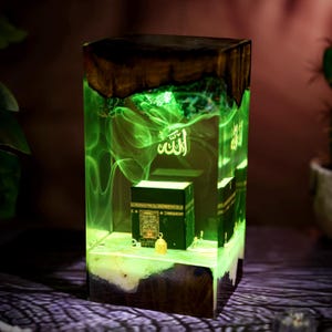 May include: A rectangular resin lamp featuring a dark wood base and top, enclosing a miniature black Kaaba model. The interior glows with a bright green light, displaying swirling smoke-like patterns and the Arabic word Allah in white script.