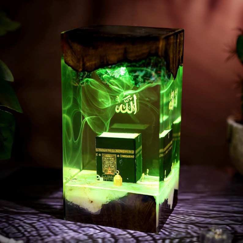 May include: A rectangular resin lamp featuring a dark wood base and top, enclosing a miniature black Kaaba model. The interior glows with a bright green light, displaying swirling smoke-like patterns and the Arabic word Allah in white script.