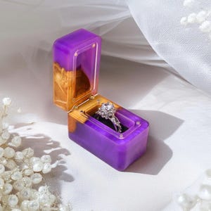Puede incluir: Caja rectangular para anillos de resina morada y dorada, abierta para mostrar un anillo de compromiso de diamantes. El interior es de terciopelo negro. El anillo tiene un diamante redondo. La caja mide unos 7 cm de largo.