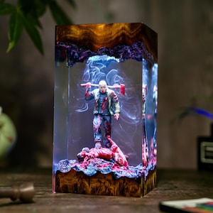 May include: A rectangular resin sculpture featuring a figure with a hockey mask holding an axe. The sculpture has a wooden base and top, with a smoky, blue and purple interior. The figure stands on a pile of red material.