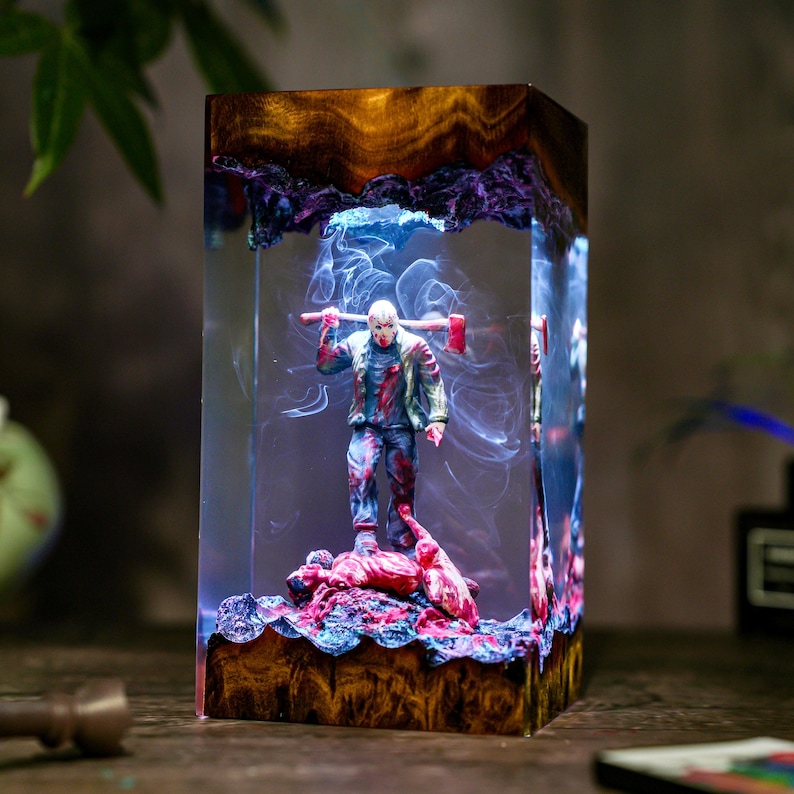 May include: A rectangular resin sculpture featuring a figure with a hockey mask holding an axe. The sculpture has a wooden base and top, with a smoky, blue and purple interior. The figure stands on a pile of red material.