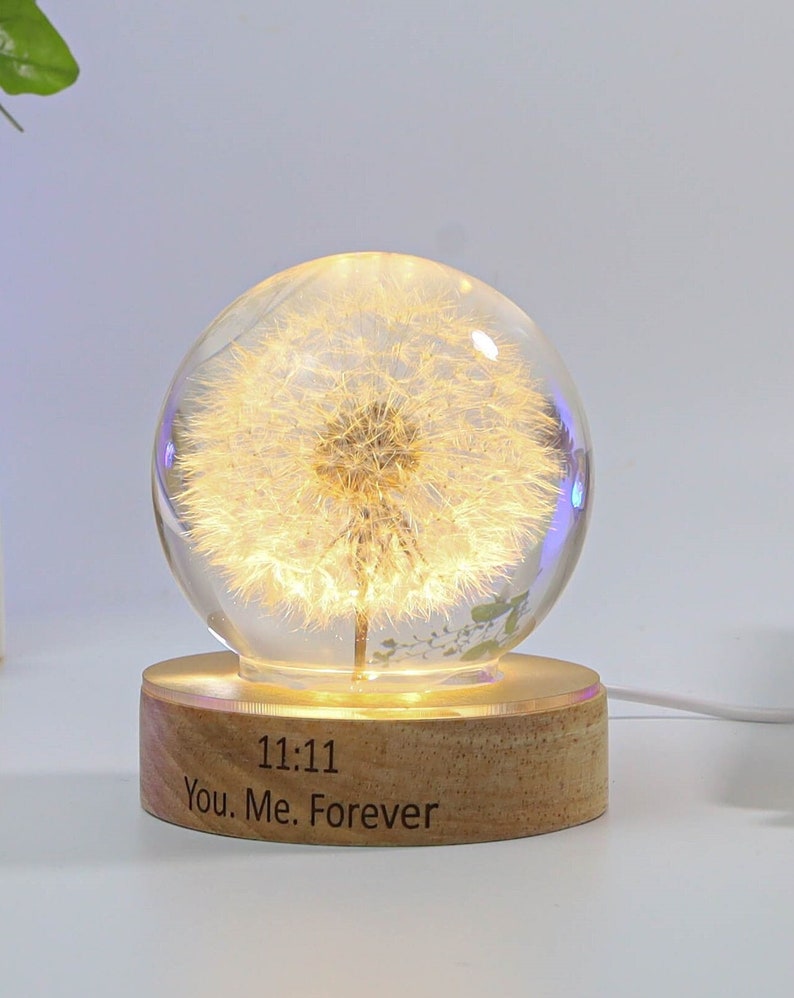 May include: A glass globe with a white dandelion inside, illuminated from within. The globe sits on a wooden base with the inscription "11:11 You. Me. Forever".