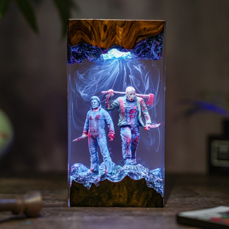 May include: A rectangular resin art piece featuring two horror movie characters, one with a hockey mask and the other in blue overalls, both holding weapons. The figures are set against a smoky, blue-lit background, encased in a wooden-edged resin block.