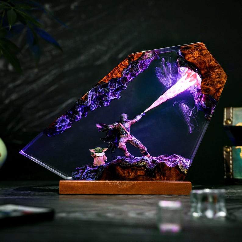 May include: A decorative lamp featuring a resin and wood frame with a scene of a armored figure in a cape firing a bright purple energy beam, accompanied by a small green creature in a tan robe, all set on a rectangular wooden base.