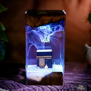 May include: A rectangular resin lamp featuring a dark wood base and top, enclosing a miniature black Kaaba model. The clear resin body contains swirling white smoke-like patterns and the Arabic word Allah illuminated in bright white light.
