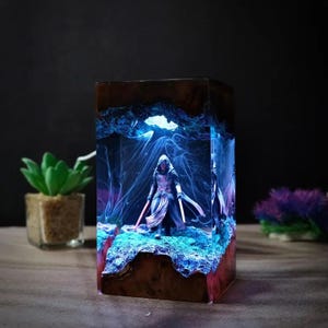 May include: A rectangular resin lamp featuring a dark wood base and top, enclosing a miniature hooded figure holding two glowing lightsabers. The interior displays swirling blue smoke-like patterns and a textured, illuminated rocky terrain.