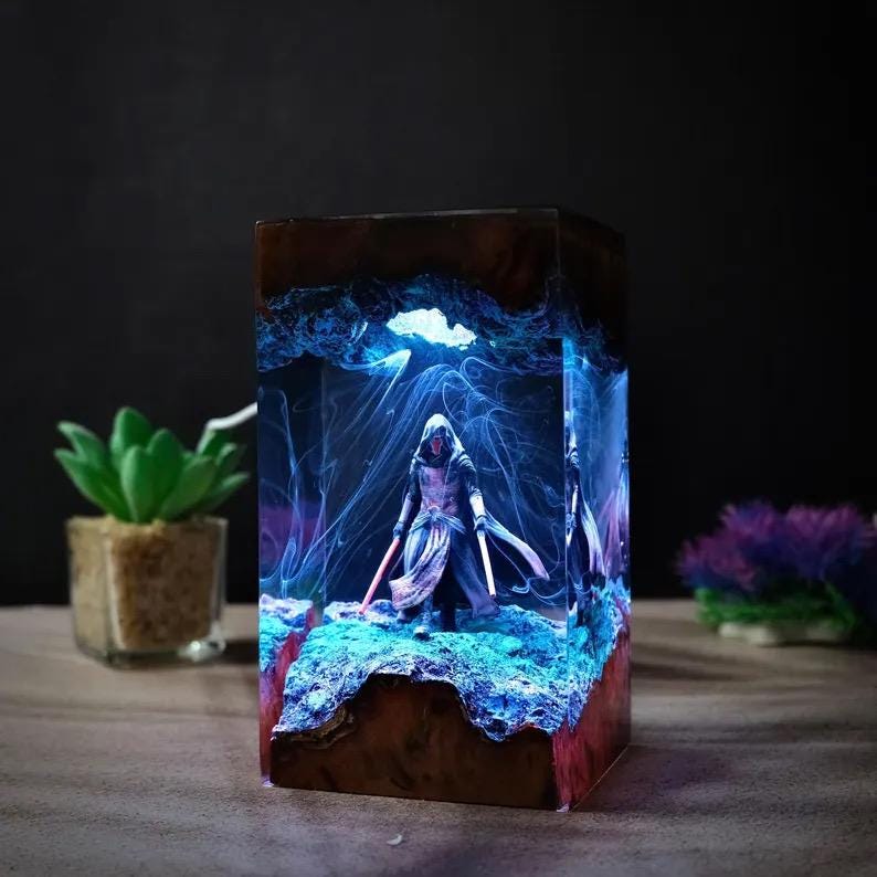 May include: A rectangular resin lamp featuring a dark wood base and top, enclosing a miniature hooded figure holding two glowing lightsabers. The interior displays swirling blue smoke-like patterns and a textured, illuminated rocky terrain.