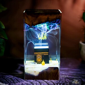 May include: A rectangular resin lamp featuring a dark wood base and top, enclosing a miniature black Kaaba model. The clear resin body contains swirling white smoke effects and a glowing gold Arabic calligraphy script floating above the structure.