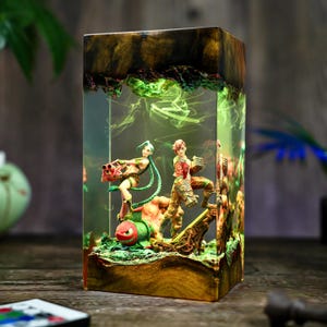 May include: A rectangular resin lamp featuring figures from a video game. The lamp has a wooden base and top, with the figures and green smoke-like effects encased in clear resin. The figures are in action poses, with one holding a large weapon.