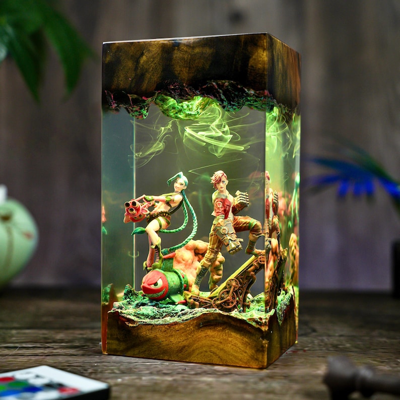 May include: A rectangular resin lamp featuring figures from a video game. The lamp has a wooden base and top, with the figures and green smoke-like effects encased in clear resin. The figures are in action poses, with one holding a large weapon.