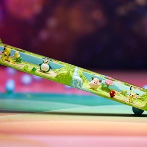 May include: A clear resin tube with a miniature landscape scene inside. The scene features a small waterfall, green trees, pink flowers, and miniature figurines of cartoon characters.
