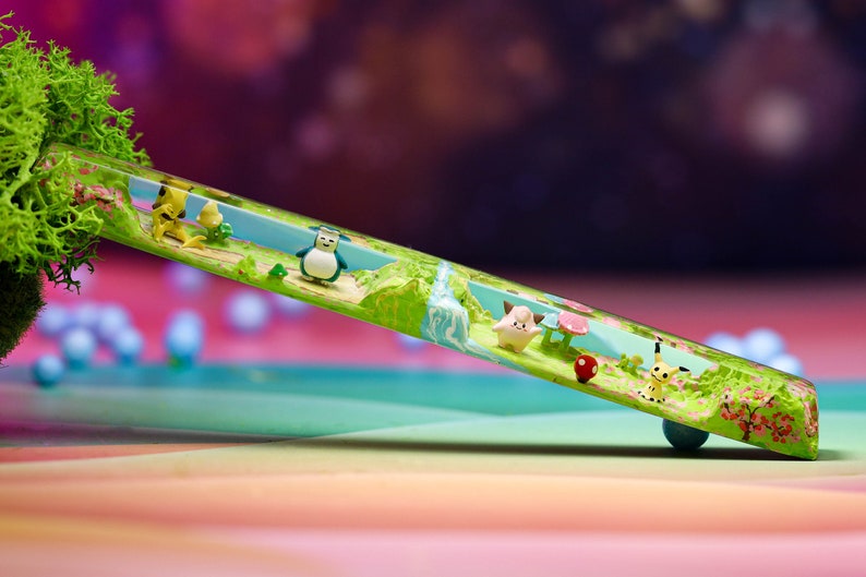 May include: A clear resin tube with a miniature landscape scene inside. The scene features a small waterfall, green trees, pink flowers, and miniature figurines of cartoon characters.
