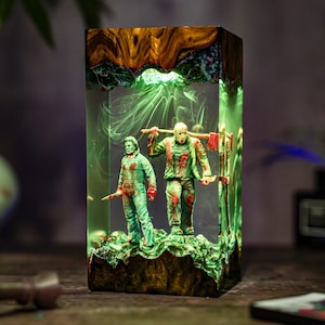 May include: A rectangular resin lamp featuring two horror movie figures. The figures are green and red, standing on a green base. The lamp has a wooden top and base, with green lighting effects throughout.