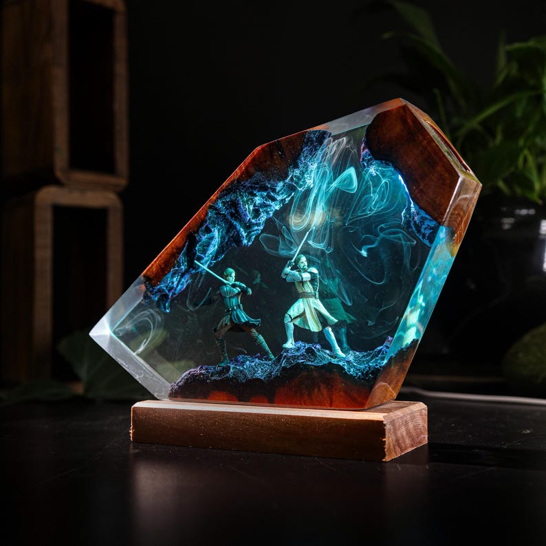 May include: A unique resin sculpture featuring two figures in a lightsaber duel, set within a clear, angled block. The figures are surrounded by blue and white swirling patterns, with a wooden base and accents. The sculpture is a decorative item.