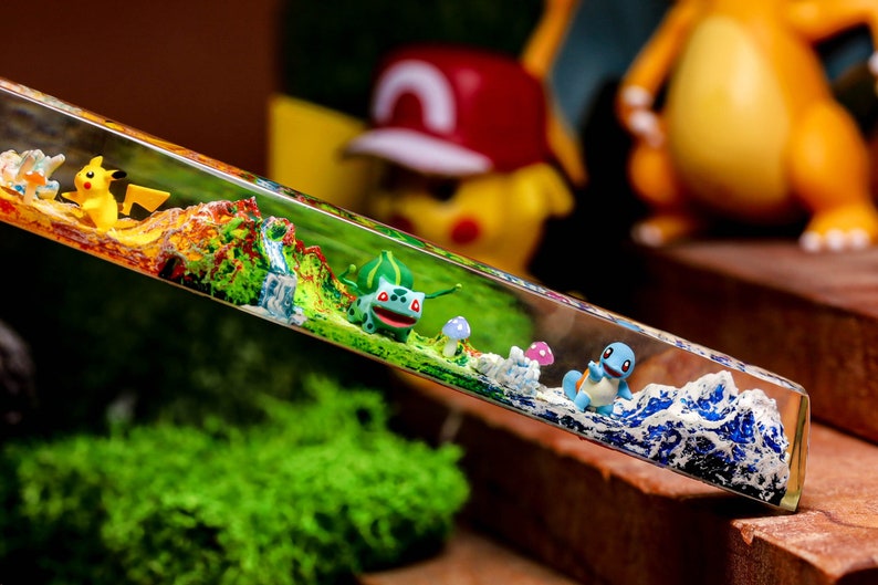 May include: A clear resin rectangular block with a miniature green and yellow Pokemon character, a blue Pokemon character, and a small mushroom. The block is on a brown surface with green moss in the background.