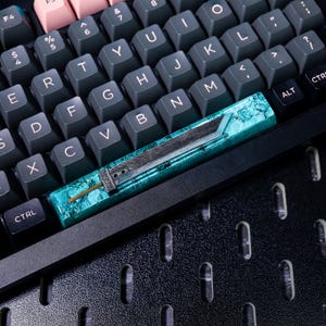 May include: A teal and silver keyboard keycap with a sword design. The keycap is on a black keyboard with a textured surface.