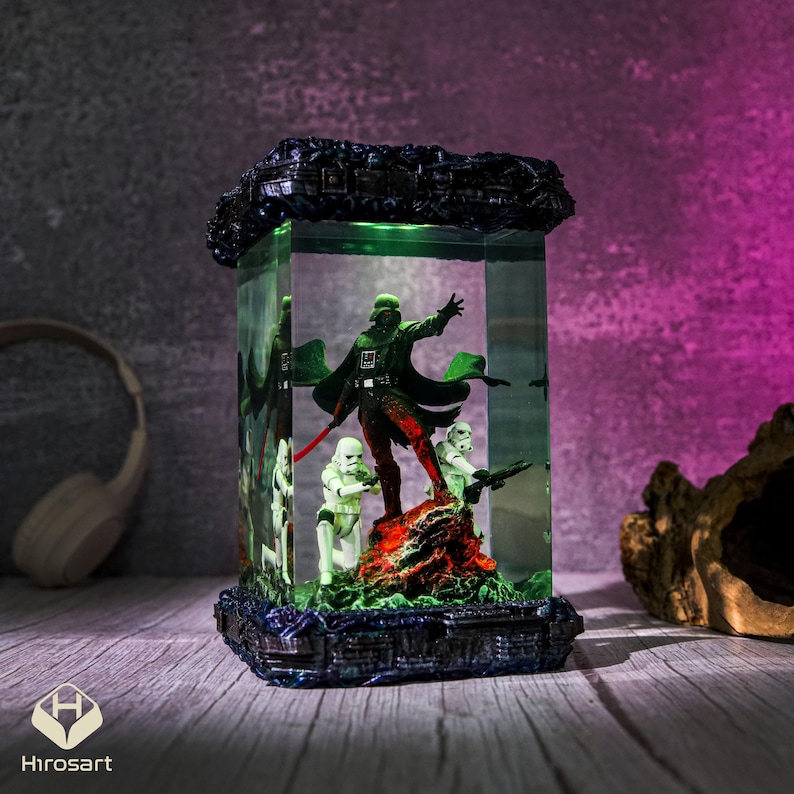 K&ouml;nnte beinhalten: Eine schwarze und gr&uuml;ne Darth Vader Figur mit einem roten Lichtschwert, die in einem klaren Glasw&uuml;rfel mit einem gr&uuml;nen Licht im Inneren steht. Der W&uuml;rfel ist von einem schwarzen Sockel mit silbernem Rand umgeben. Zwei wei&szlig;e Stormtrooper Figuren stehen hinter Darth Vader.