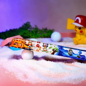 May include: A clear glass tube with a colorful scene inside. The scene includes a campfire, a boat, and cartoon characters. The tube is resting on a white surface.