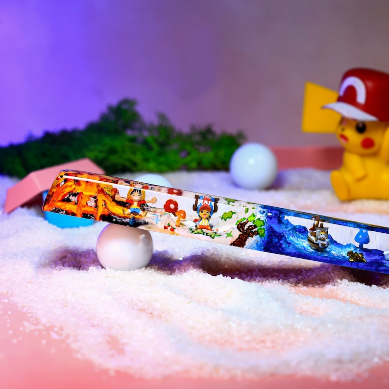 May include: A clear glass tube with a colorful scene inside. The scene includes a campfire, a boat, and cartoon characters. The tube is resting on a white surface.