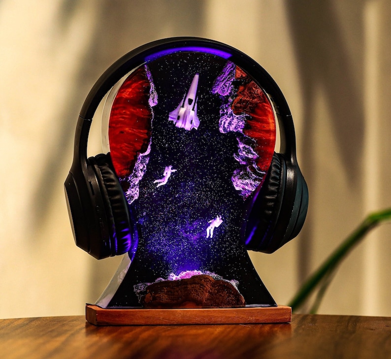 May include: A black headphone stand with a resin base featuring a galaxy scene with a spaceship and two astronauts. The base is lit with purple and blue LED lights.