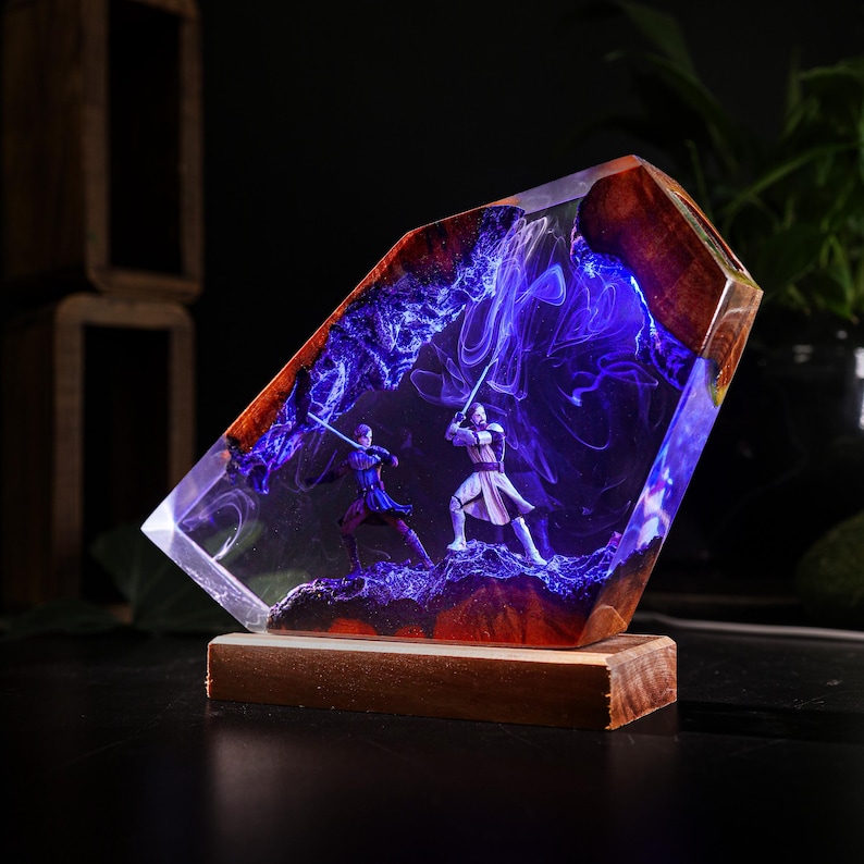 May include: A resin sculpture featuring two figures with lightsabers in a battle scene. The sculpture is encased in clear resin with a blue and purple nebula effect, set on a wooden base. The edges are framed with wood.