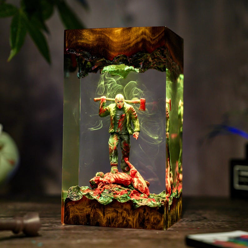 May include: A rectangular resin sculpture featuring a figure with a red and green outfit holding an axe. The figure stands on a base with red and green details, encased in clear resin with a wood-grain top.
