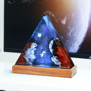 May include: A triangular resin lamp with a blue and purple galaxy design, a white silhouette of a flying animal, and a wooden base.