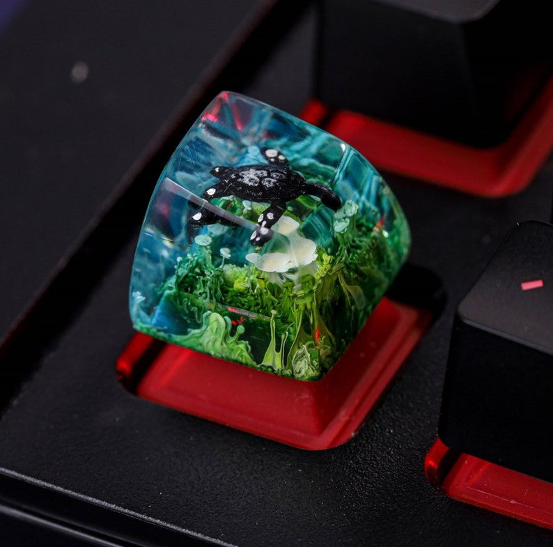 May include: A black turtle figurine encased in clear resin with green and blue swirls. The resin keycap is a decorative accent for a keyboard.