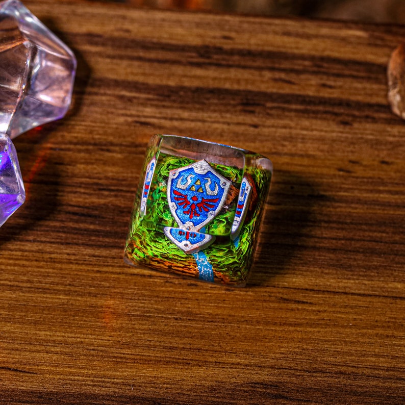 May include: A clear resin keycap with a green, blue, and red shield design. The shield has a stylized Triforce symbol and is surrounded by a green and blue background.