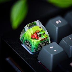 May include: A miniature house with a red roof and green grass is encased in a clear resin keycap. The keycap is sitting on a black keyboard.